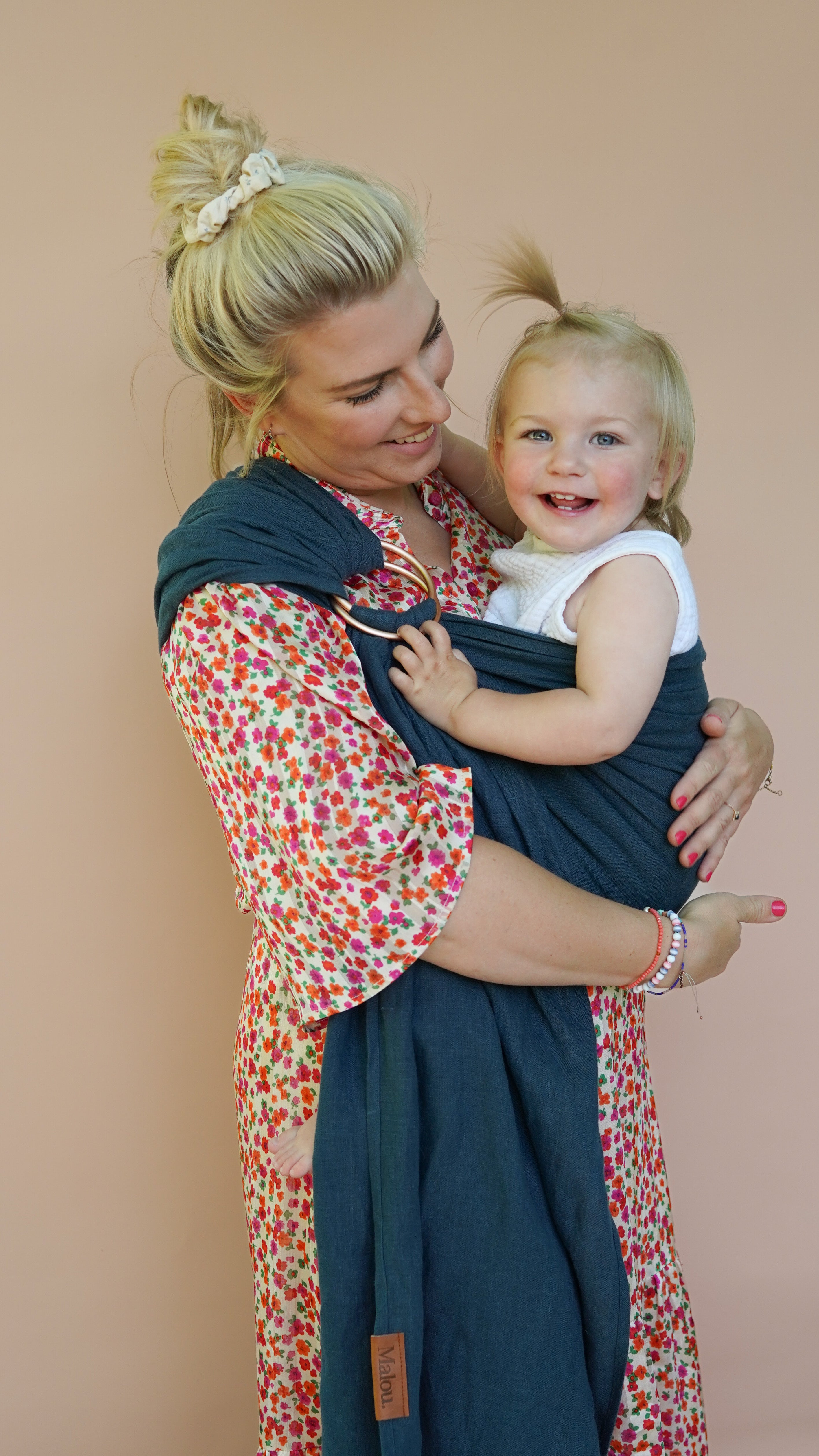 Blonde Frau mit ihrem Baby in einer dunkelblau-grünen Ringsling. Das Kind schaut in die Kamera, während die Mutter es glücklich ansieht.