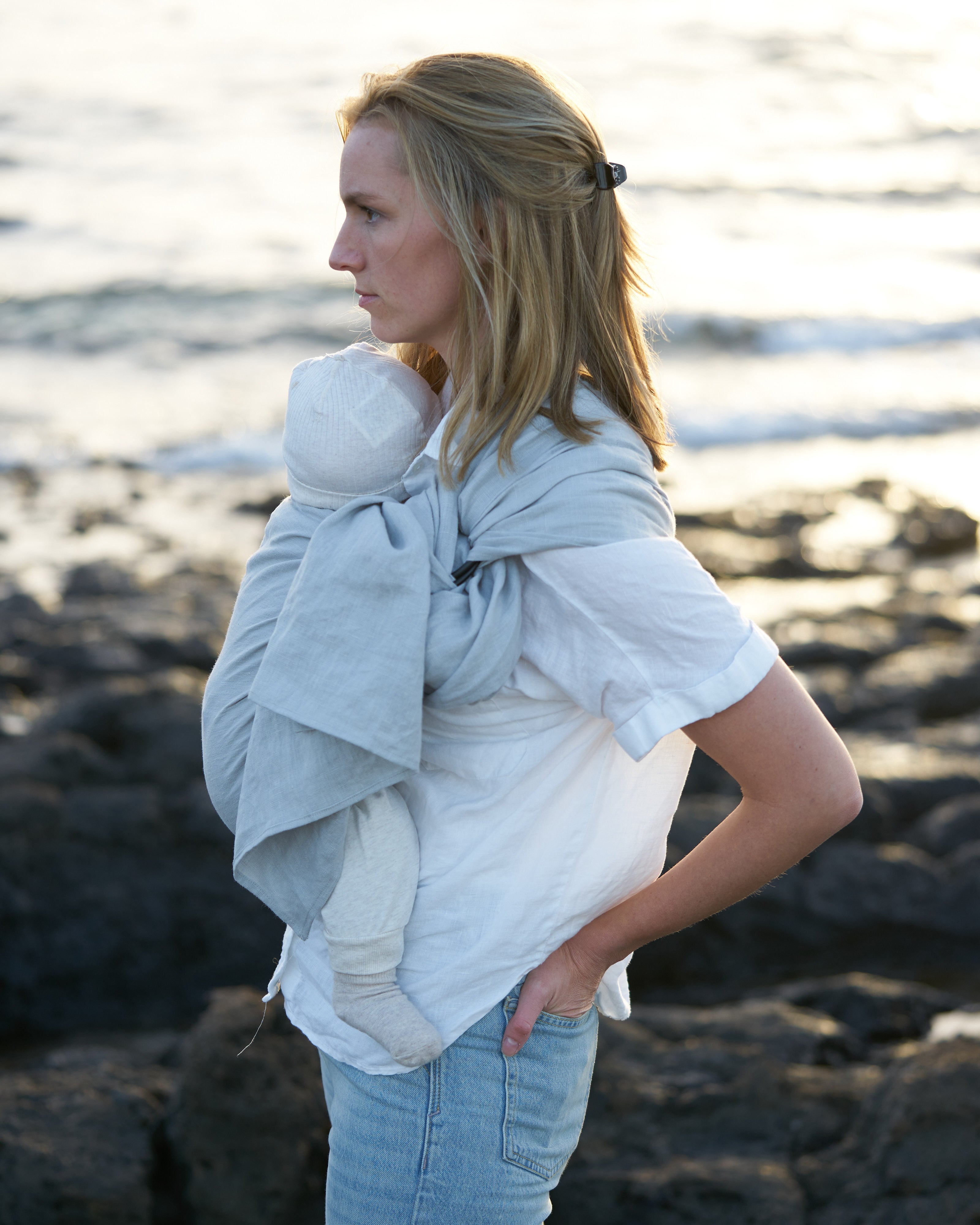 Eine blonde Frau mit Baby in einer grauen Ring Sling steht am Meer. 